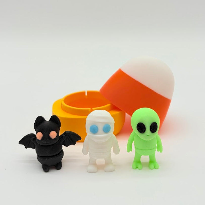 Three small figurines (bat, ghost, alien) in front of a candy corn-shaped container on a white background.